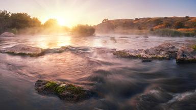 Sonnenaufgang über einem Fluss mit nebliger Wasseroberfläche und feuchtem Uferbewuchs. Felsen liegen im Wasser.